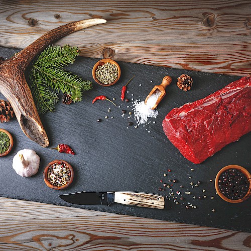 On a slate board: a large raw steak, a deer antler, small bowls with spices, garlic, chili, pine cones, and a knife, all arranged for a rustic meat prep scene.