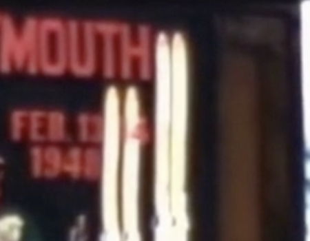 A blurry sign reading “DARTMOUTH” in red letters, with a person portrait and some tall white shapes (possibly sails or lights) beside it.