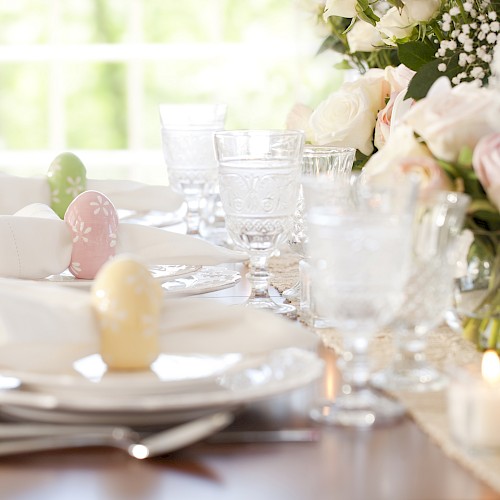 A beautifully set Easter table with pastel eggs, floral arrangements, wine glasses, and a candle in natural light.