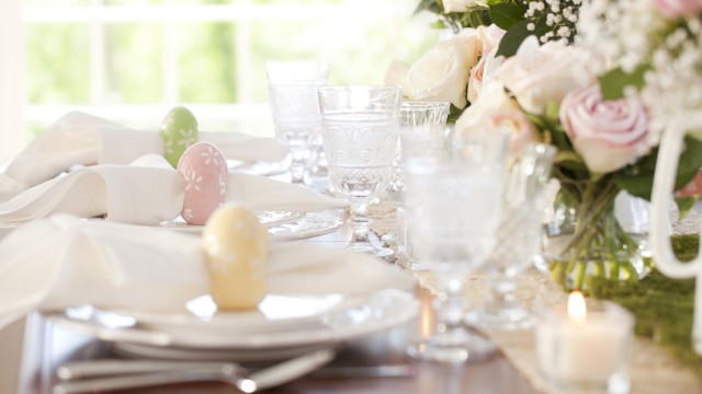 A beautifully set Easter table with pastel eggs, floral arrangements, wine glasses, and a candle in natural light.