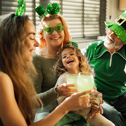 A family celebrates St. Patrick's Day with green accessories, laughing, and enjoying drinks inside a cozy home.