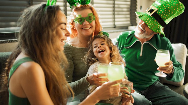 A family celebrates St. Patrick's Day with green accessories, laughing, and enjoying drinks inside a cozy home.