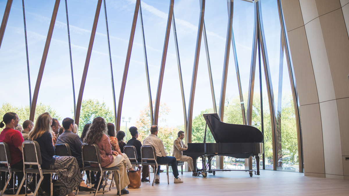 An audience watches a pianist perform in a modern concert hall with large glass windows and scenic outdoor view.