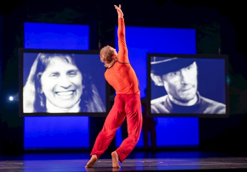A dancer in an orange top and red pants performs on stage with video screens of faces behind her.