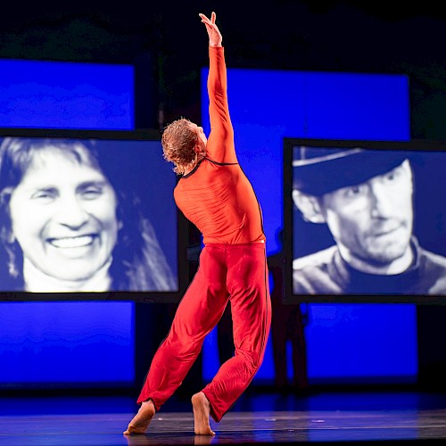 A dancer in an orange top and red pants performs on stage with video screens of faces behind her.