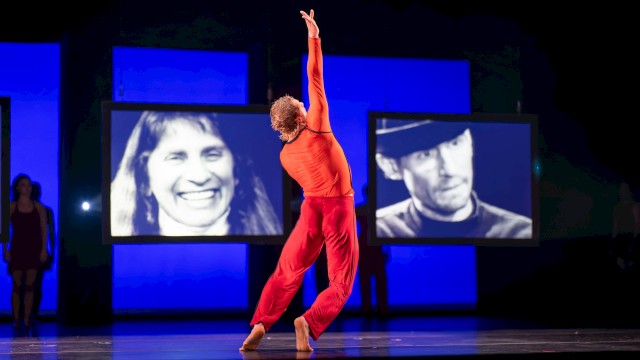 A dancer in an orange top and red pants performs on stage with video screens of faces behind her.