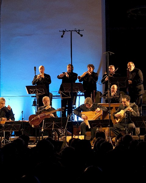 An orchestra performs live on stage with blue lighting, playing various instruments in front of an audience.