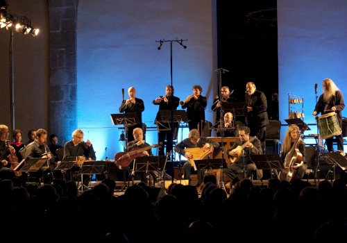 An orchestra performs live on stage with blue lighting, playing various instruments in front of an audience.