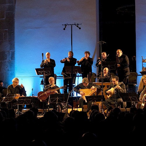 An orchestra performs live on stage with blue lighting, playing various instruments in front of an audience.