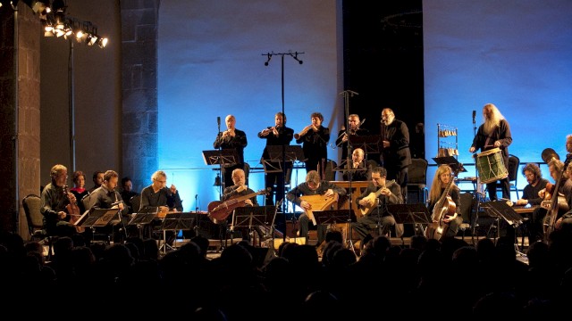 An orchestra performs live on stage with blue lighting, playing various instruments in front of an audience.