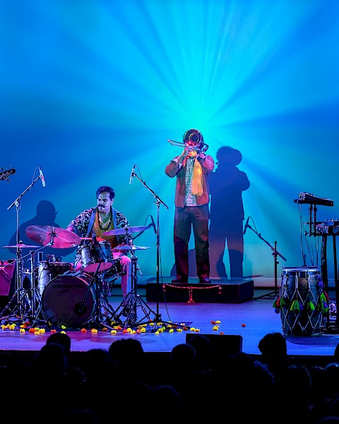 A band performs on stage with colorful lighting, including drums, keyboard, guitars, and wind instruments, for an audience.
