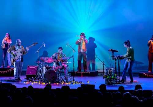 A band performs on stage with colorful lighting, including drums, keyboard, guitars, and wind instruments, for an audience.