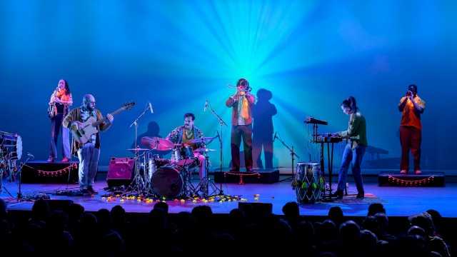 A band performs on stage with colorful lighting, including drums, keyboard, guitars, and wind instruments, for an audience.