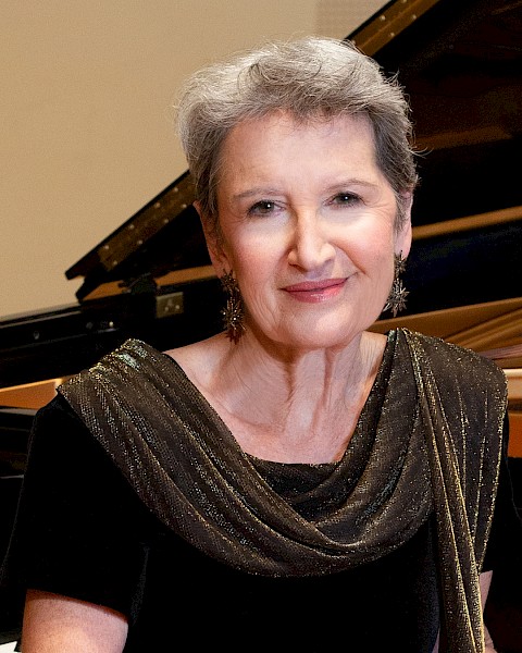 An elegant woman with short gray hair, wearing a black dress with a shimmering scarf, sits beside a grand piano in a music room.