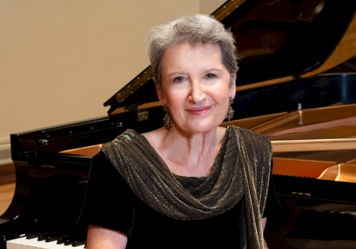 An elegant woman with short gray hair, wearing a black dress with a shimmering scarf, sits beside a grand piano in a music room.