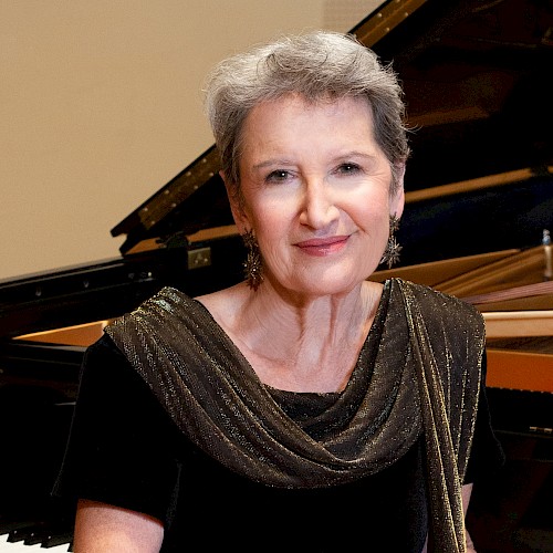 An elegant woman with short gray hair, wearing a black dress with a shimmering scarf, sits beside a grand piano in a music room.