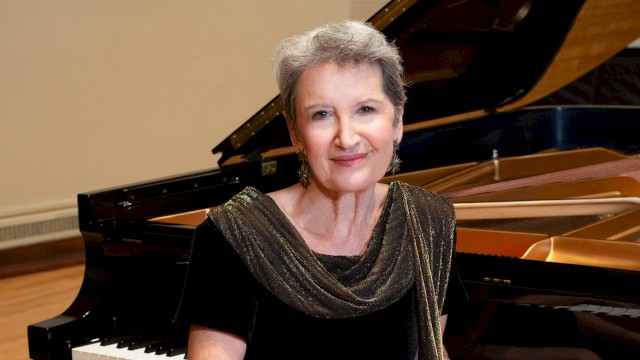 An elegant woman with short gray hair, wearing a black dress with a shimmering scarf, sits beside a grand piano in a music room.