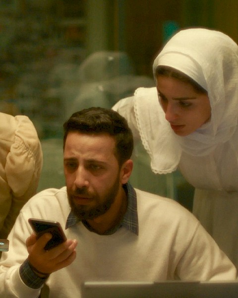 Four people, including a man with a beard, look at a phone and a laptop with concern inside a room.