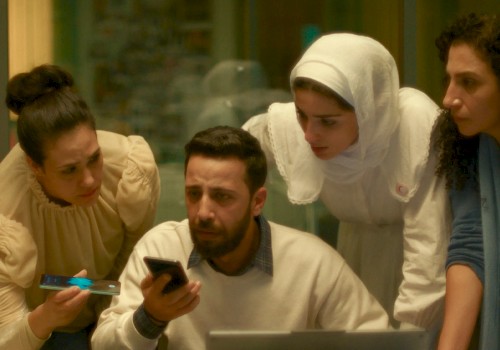 Four people, including a man with a beard, look at a phone and a laptop with concern inside a room.