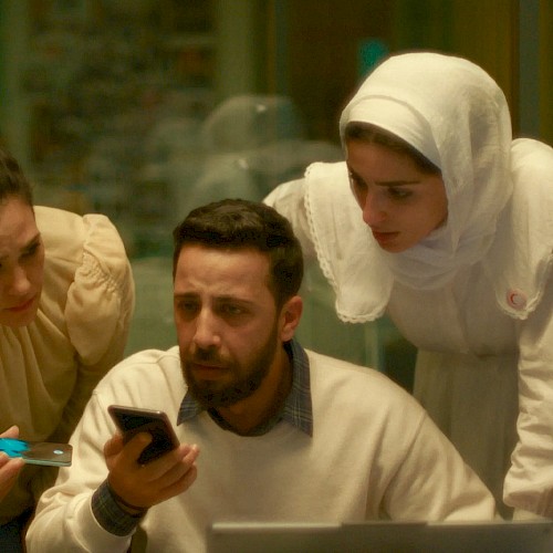 Four people, including a man with a beard, look at a phone and a laptop with concern inside a room.