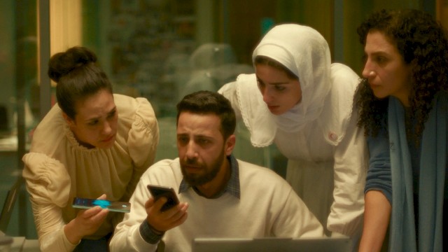 Four people, including a man with a beard, look at a phone and a laptop with concern inside a room.