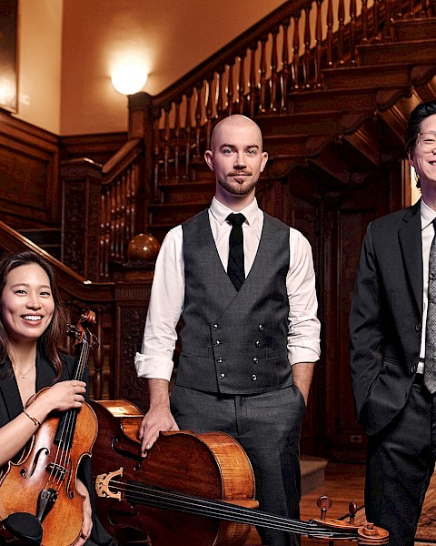 A group of five musicians dressed in formal attire with violins, a cello, and a viola, standing inside a richly wood-paneled building.