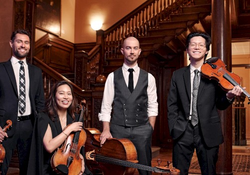 A group of five musicians dressed in formal attire with violins, a cello, and a viola, standing inside a richly wood-paneled building.