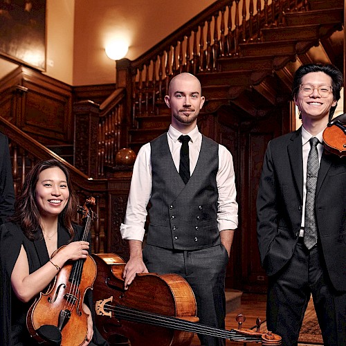 A group of five musicians dressed in formal attire with violins, a cello, and a viola, standing inside a richly wood-paneled building.