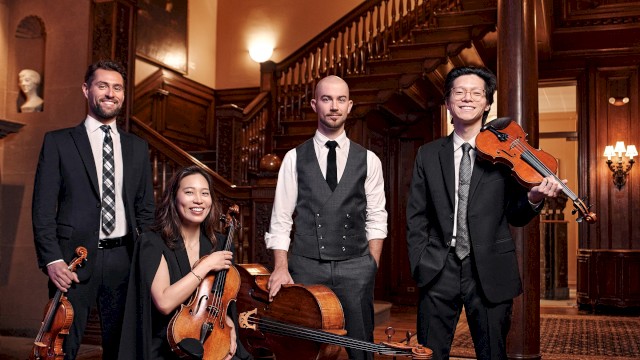 A group of five musicians dressed in formal attire with violins, a cello, and a viola, standing inside a richly wood-paneled building.