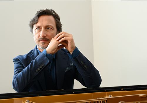 A man with gray hair and a beard, wearing a blue jacket, is sitting at a piano and looking thoughtfully to the side.