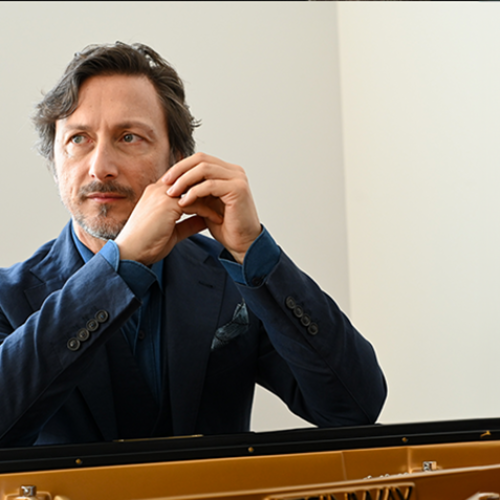 A man with gray hair and a beard, wearing a blue jacket, is sitting at a piano and looking thoughtfully to the side.