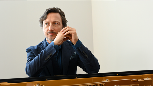 A man with gray hair and a beard, wearing a blue jacket, is sitting at a piano and looking thoughtfully to the side.