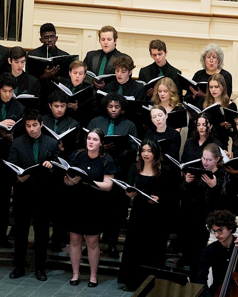 A choir of diverse young singers performs in a church or concert hall, accompanied by a cellist, under the direction of a conductor.