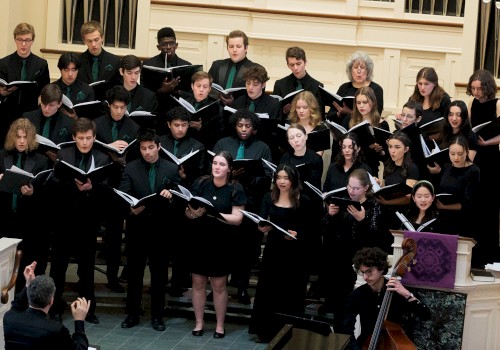 A choir of diverse young singers performs in a church or concert hall, accompanied by a cellist, under the direction of a conductor.