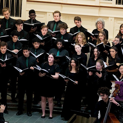 A choir of diverse young singers performs in a church or concert hall, accompanied by a cellist, under the direction of a conductor.