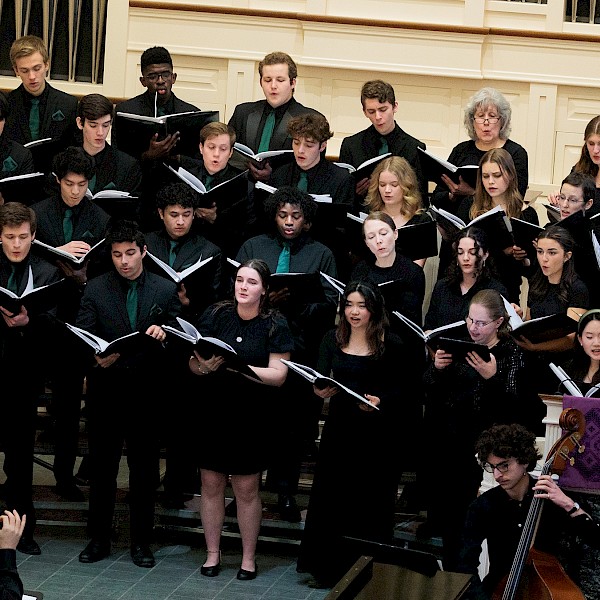A choir of diverse young singers performs in a church or concert hall, accompanied by a cellist, under the direction of a conductor.