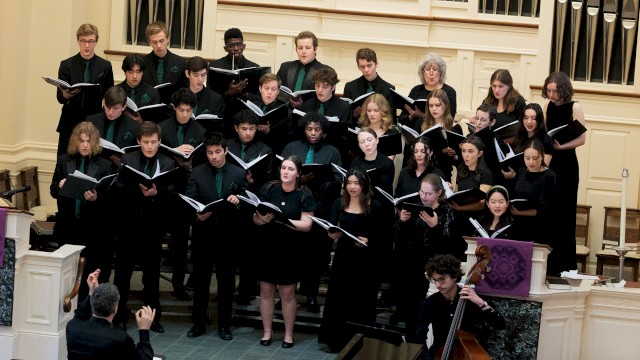 A choir of diverse young singers performs in a church or concert hall, accompanied by a cellist, under the direction of a conductor.