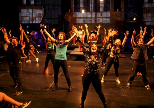 A group of diverse performers dance enthusiastically on stage during a lively theatrical production with an audience watching.