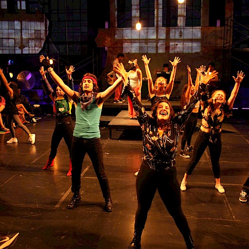 A group of diverse performers dance enthusiastically on stage during a lively theatrical production with an audience watching.