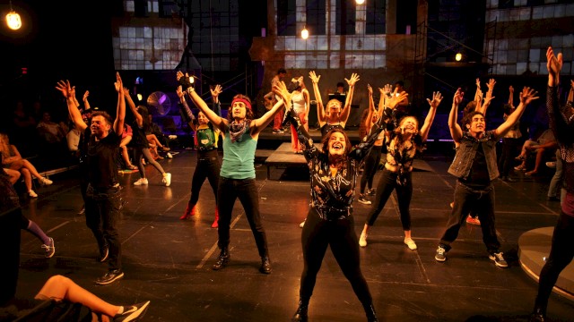 A group of diverse performers dance enthusiastically on stage during a lively theatrical production with an audience watching.