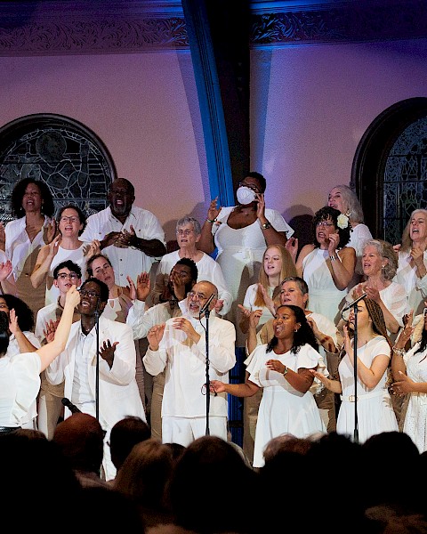 A diverse choir dressed in white performs passionately on stage in front of an audience, with stained glass windows in the background.