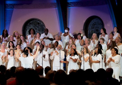 A diverse choir dressed in white performs passionately on stage in front of an audience, with stained glass windows in the background.