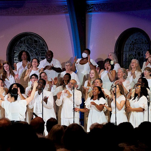 A diverse choir dressed in white performs passionately on stage in front of an audience, with stained glass windows in the background.