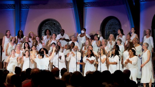 A diverse choir dressed in white performs passionately on stage in front of an audience, with stained glass windows in the background.