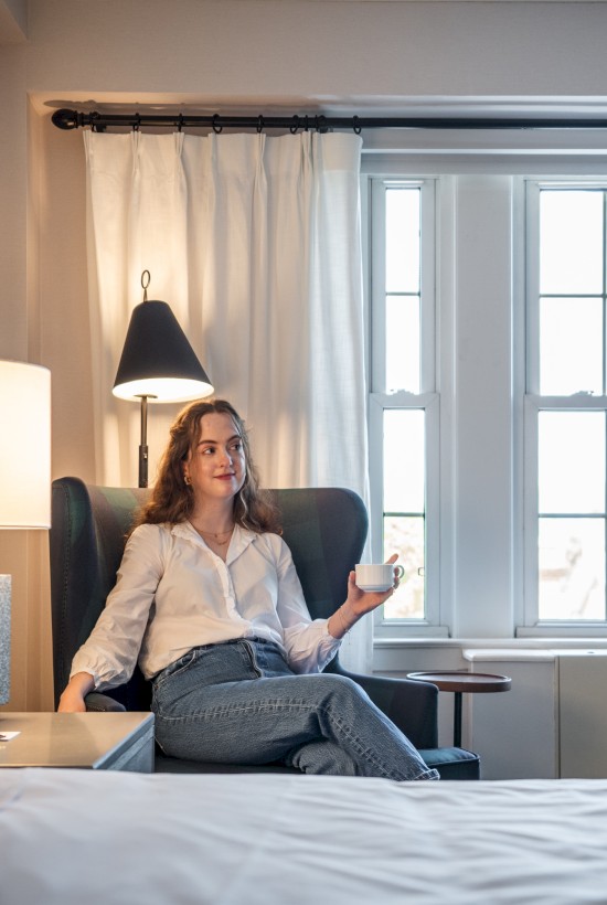 A woman relaxes in a chair, holding a cup, in a cozy room illuminated by natural light coming through large windows.