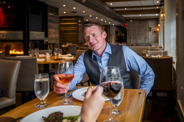 A man in a blue shirt and vest dining at a restaurant, holding a glass of rosé wine.