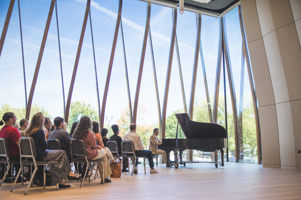 A pianist performs in front of an audience in a modern, glass-walled concert hall with a scenic outdoor view.