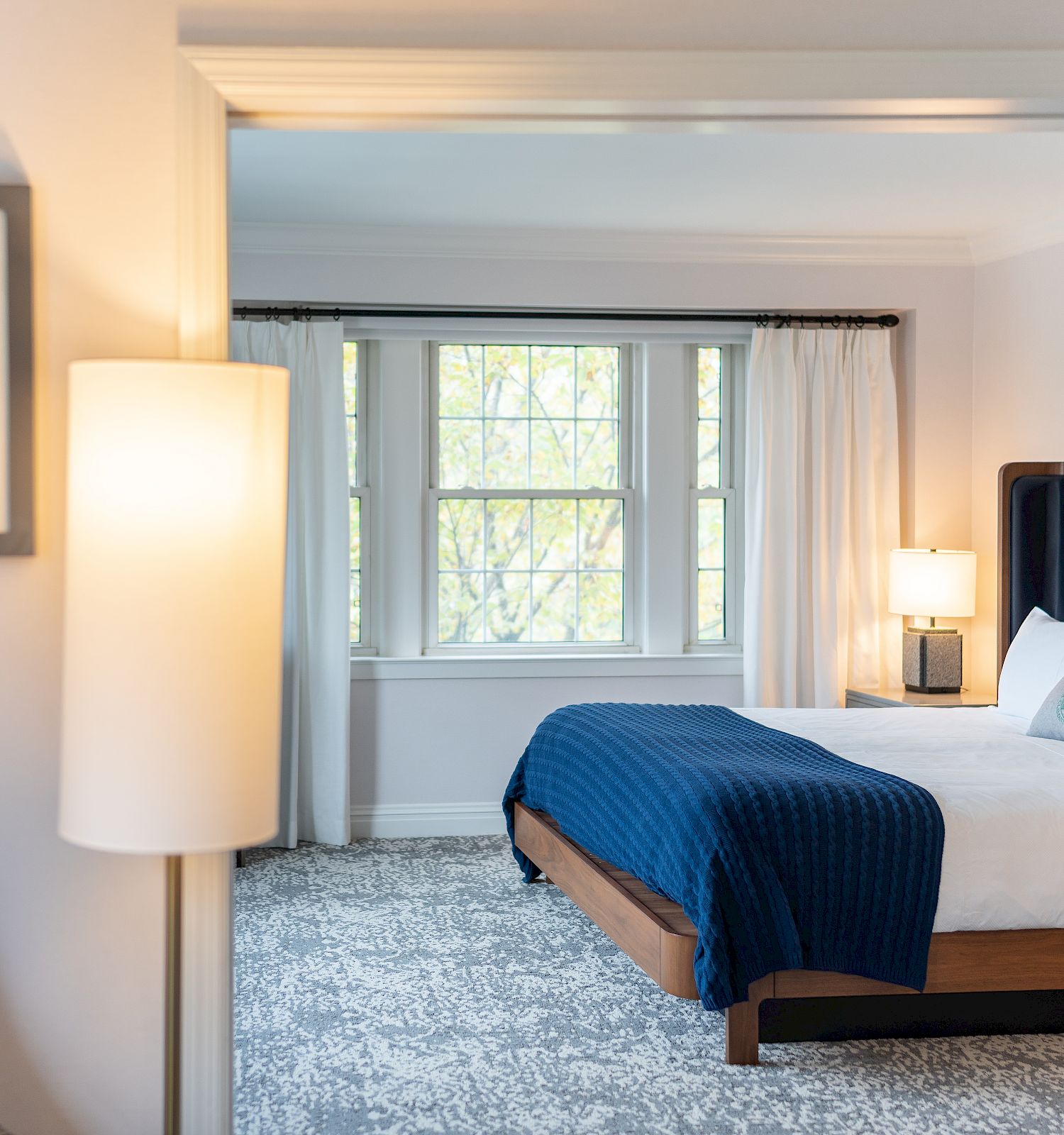 This image depicts a cozy bedroom with a neatly made bed, blue bedding, white curtains, and a framed poster on the wall near a standing lamp.