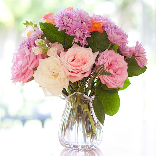 A clear vase holds a bouquet of pink and white flowers, including roses and chrysanthemums, set against a softly blurred background.