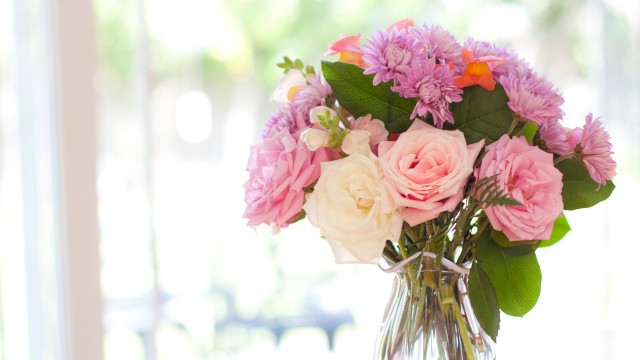 A clear vase holds a bouquet of pink and white flowers, including roses and chrysanthemums, set against a softly blurred background.